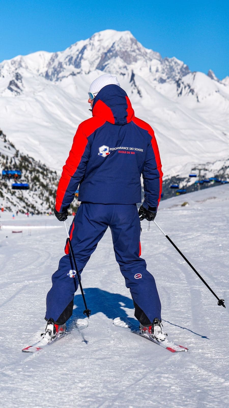A Performance Ski School instructor on skis, seen from behind, facing the snow-covered peaks